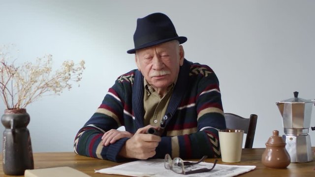 Dolly-in portrait shot of stylish elderly man in hat sitting at table and smoking pipe, then looking at camera and smiling