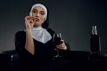 Cropped half-turn shot of a nun, sitting on a black chair. She's wearing dark nun's clothing. The nun is smoking and holding glass of wine in her hand. There is a bottle with alcohol on chair armrest. - obrazy, fototapety, plakaty