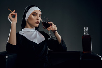 Cropped half-turn shot of a nun, sitting on a chair. She's wearing dark nun's clothing. The nun is drinking wine and holding cigarette in her hand. There is a bottle with alcohol on a chair armrest. - obrazy, fototapety, plakaty