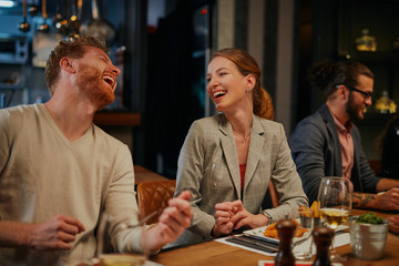 Charming smiling caucasian blonde sitting in restaurant, eating dinner and chatting with friend. In background are friends.