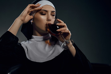 Portrait shot of a nun, sitting on a chair. She's wearing dark nun's clothing. The nun is drinking...