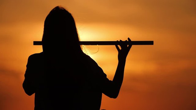 Close up silhouette of a beautiful girl with long hair playing on flute in front of sunset. Slow motion. Enlightenment