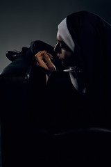 Upward half-turn back view shot of a nun, sitting on a chair. She's looking down, her left hand is...