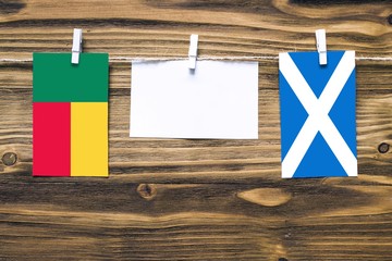 Hanging flags of Benin and Scotland attached to rope with clothes pins with copy space on white note paper on wooden background.Diplomatic relations between countries.