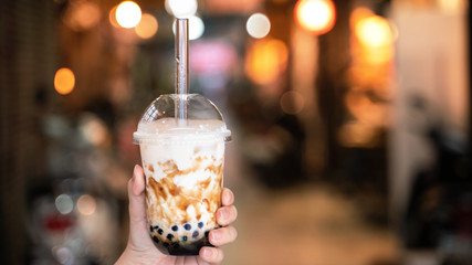 Young woman is holding, drinking brown sugar flavored tapioca pearl bubble milk tea with glass straw in night market of Taiwan, close up, bokeh