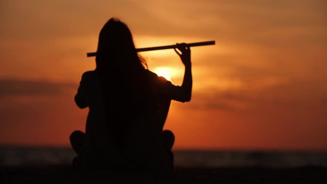 Close up silhouette of a beautiful girl with long hair playing on flute in front of sunset. Slow motion. Enlightenment
