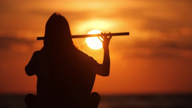 Close up silhouette of a beautiful girl with long hair playing on flute in front of sunset. Slow motion. Enlightenment