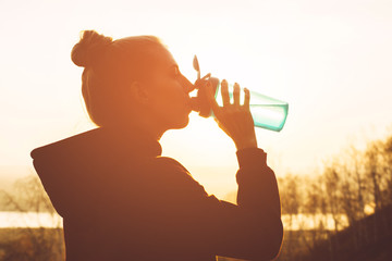 Young girl in sportswear drinks water from a shaker at sunset