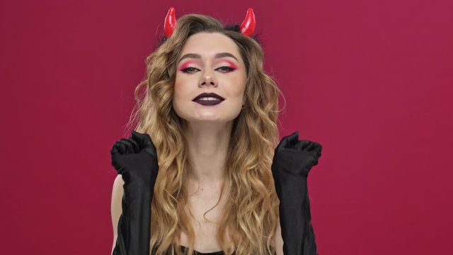 Beautiful and crazy devil woman in carnival costume flirt to the camera and bite isolated over red wall background 