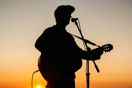 Silhouette of a man playing guitar and singing into a microphone at sunset
