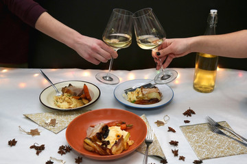 Festive holiday table with pork belly roast, mashed potatoes and roasted vegetables. Unrecognizable people cheering with white wine. 