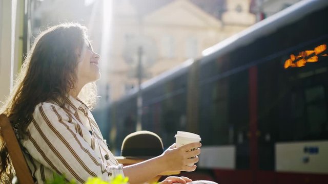 Side view elegant lady enjoying coffee in sunlight at sidewalk cafe in the center of Prague. Cropped Lonely girl drinking coffee in sunny morning, looking at empty city street and tram passing by 