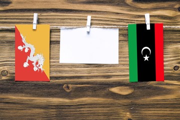 Hanging flags of Bhutan and Libya attached to rope with clothes pins with copy space on white note paper on wooden background.Diplomatic relations between countries.