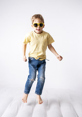 A little boy in yellow sunglasses and a yellow skirt jumps on a white mattress. Studio portrait on a white background.