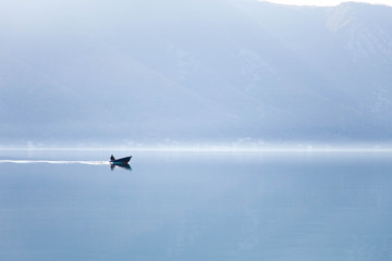 Fototapeta premium Blue nature background. Boat with fisherman on sea. Fishing in foggy morning lake. Amazing seaside landscape with mountains, silence, calmness. Reflection in still water of the Kotor Bay, Montenegro.