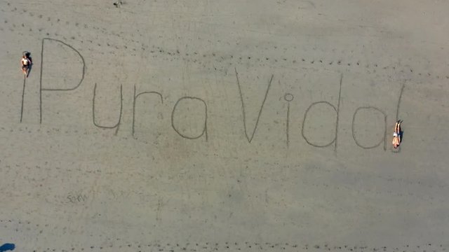 Aerial: Zooming Out at Sand Writing Saying "Pura Vida!" and Two People Lying on the Exclamation Marks - Playa Espadilla, Costa Rica