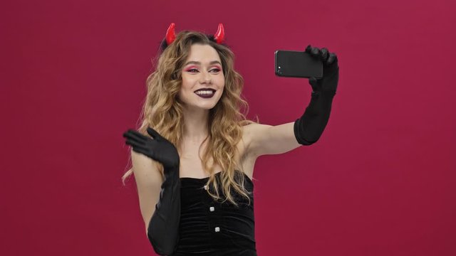 Beautiful and crazy devil woman in carnival costume is making selfie on smartphone isolated over red wall background