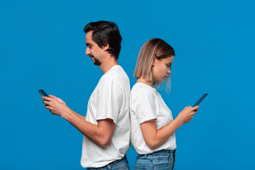 Blond woman and brunet man in white tess and blue jeans looking at screens of their mobile phones standing with their back to each other.