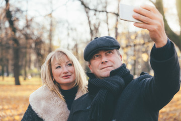 Fototapeta premium Happy blonde mature woman and beautiful middle-aged brunette take selfie on mobile phone. Loving couple of 45-50 years old walks in autumn park in warm clothes, in coat and enjoys life