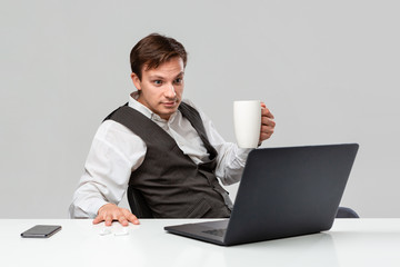 Businessman in a white t-shirt and grey vest is shocked checking information on laptop sitting at the white table drinking coffee.