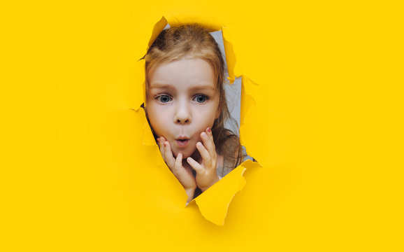 Naklejki Funny red-haired child girl peeping through hole on yellow paper. The concept of surprise, joyful mood from what he saw. Discounts, sales, surprise. Copy space.