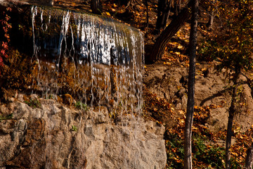 Waterfall from stones in the park. Against the background of autumn foliage and trees of orange and yellow.