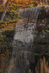 Waterfall from stones in the park. Against the background of autumn foliage and trees of orange and yellow.