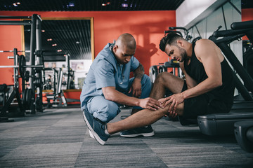 Fototapeta na wymiar attentive african american doctor examining injured knee of sportsman suffering from pain