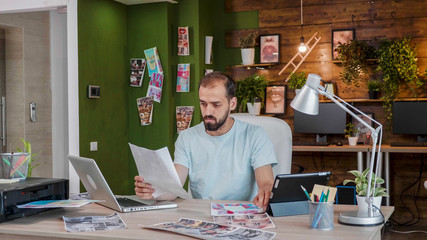 Young designer looking at a sheet holding it in his hand while sitting at the table in front of a laptop