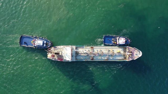 Aerial drone video of tug boats manoeuvring industrial fuel and gas carrier tanker by pushing or pulling in Mediterranean shipyard