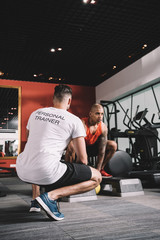 Obraz premium back view of personal trainer supervising african american athlete lifting weight in gym