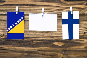 Hanging flags of Bosnia Herzegovina and Finland attached to rope with clothes pins with copy space on white note paper on wooden background.Diplomatic relations between countries.