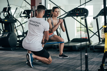 back view of personal trainer controlling attractive sportswoman exercising with resistance band - obrazy, fototapety, plakaty