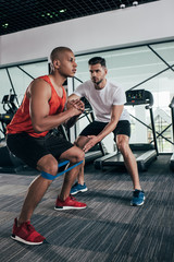 attentive trainer supporting african american sportsman warming up in gym