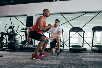 attentive trainer looking at young african american sportsman warming up in gym - Powered by Adobe