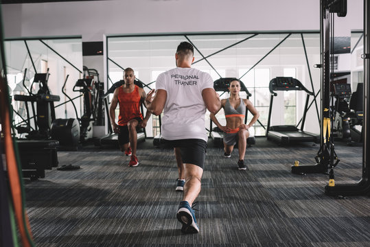 back view of personal trainer doing lunges exercise together with multicultural athletes in gym