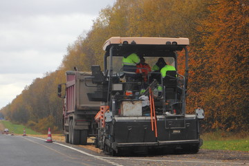 Road works, asphalt paver and dump truck on country roadside on autumn day