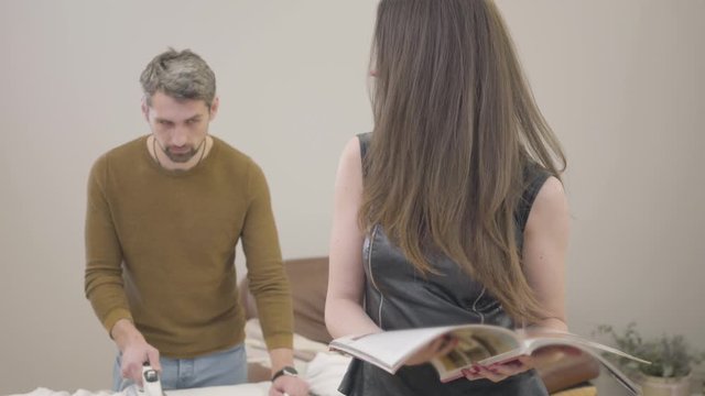 Adult handsome Caucasian man ironing at the background as young brunette woman reading the magazine. Guilty husband or henpecked doing housework while his wife resting at home.