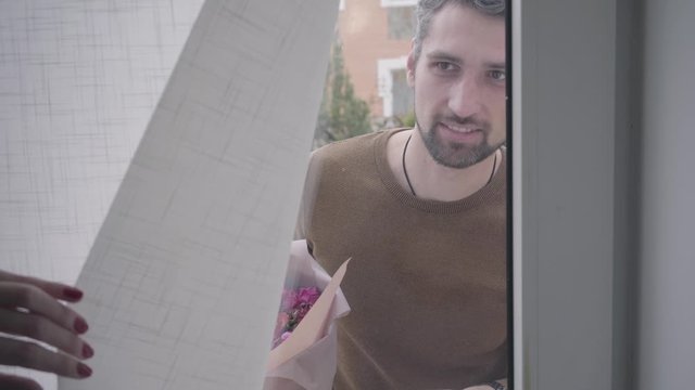 Portrait of an adult Caucasian gray-haired man standing behind the glass door with a bouquet of violet roses and asking to open. Young female hand holding the curtain and clothing it at the end.