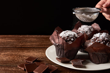 Still life on old wooden board table with three cocoa muffins with sugar and pieces of chocolate. Female hand holding strainer on black background.