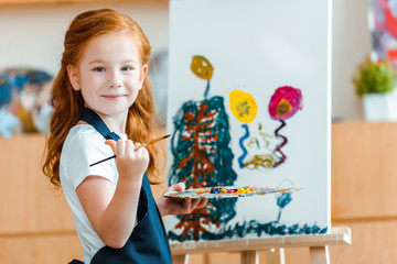 smiling redhead child standing near painting on canvas in art school © LIGHTFIELD STUDIOS