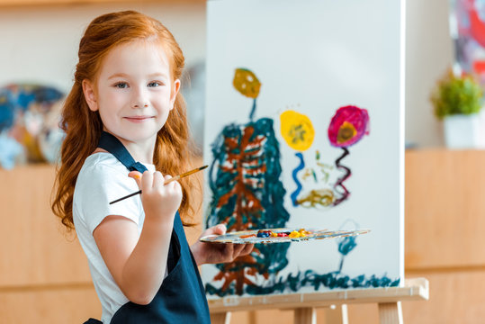 smiling redhead child standing near painting on canvas in art school