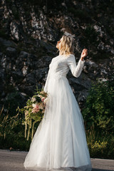 Fototapeta na wymiar Beautiful elegant bride in lace wedding dress with long full skirt and long sleeves. She is holding a big bouquet of flowers. Rock in the background.