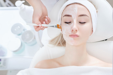 Close-up of hands of cosmetologist woman applying healing cream with brush on patient's face to young pretty caucasian woman lying on couch in beauty salon. Concept of antiaging facial skin treatments