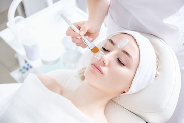 Naklejka premium Close-up of hands of cosmetologist woman applying healing cream with brush on patient's face to young pretty caucasian woman lying on couch in beauty salon. Concept of antiaging facial skin treatments