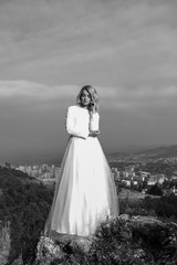 Fototapeta na wymiar Beautiful elegant bride in lace wedding dress with long full skirt and long sleeves. Pretty girl in white. Nature, with city in the background. Black and white photo.