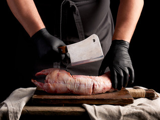 chef in black latex gloves holds a big knife and cuts into pieces raw rabbit meat on a brown wooden...