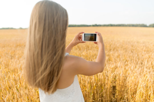 technology, summer and people concept - young girl taking picture by smartphone on cereal field