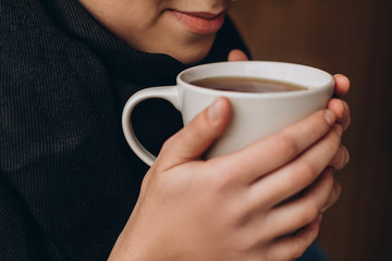 a mug of hot coffee or tea in the hands of a young pretty girl near her lips. Cropped photo with focus on cup