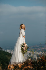 Beautiful elegant bride in lace wedding dress with long full skirt and long sleeves. She is holding a big bouquet of flowers. Nature, with city in the background.